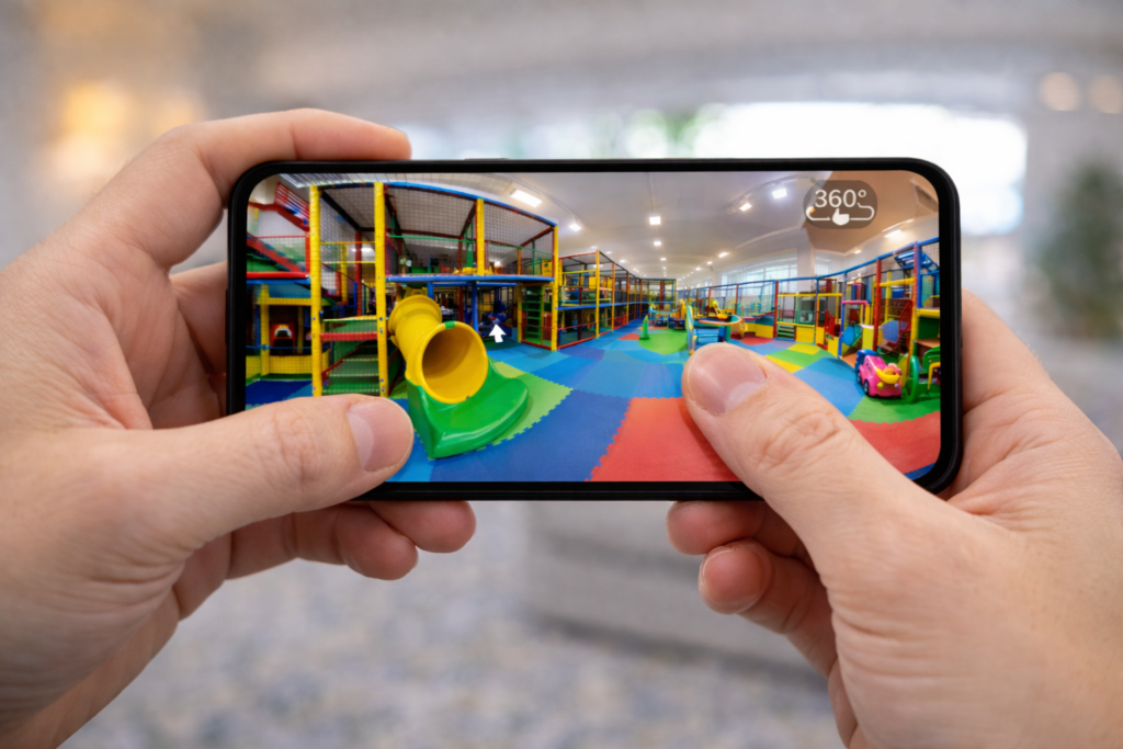 Zwei Hände halten ein Smartphone, während ein Daumen auf dem Display ein 360-Grad-Foto eines Indoor-Spielplatzes verschiebt.